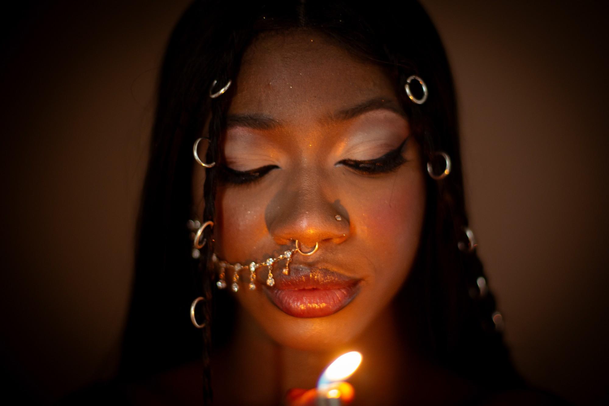 Close portrait lit by a single lighter flame: eyes lowered, silver rings in the braids, a crystal chain running from the septum ring to the ear.