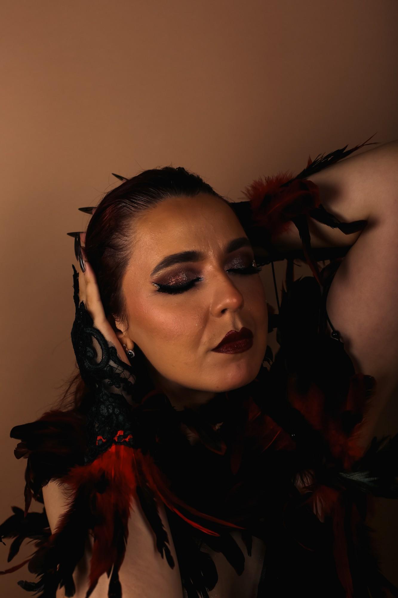 Portrait with eyes closed and one arm raised behind the head, burgundy lips and glitter liner, red and black feathers and a black lace glove.