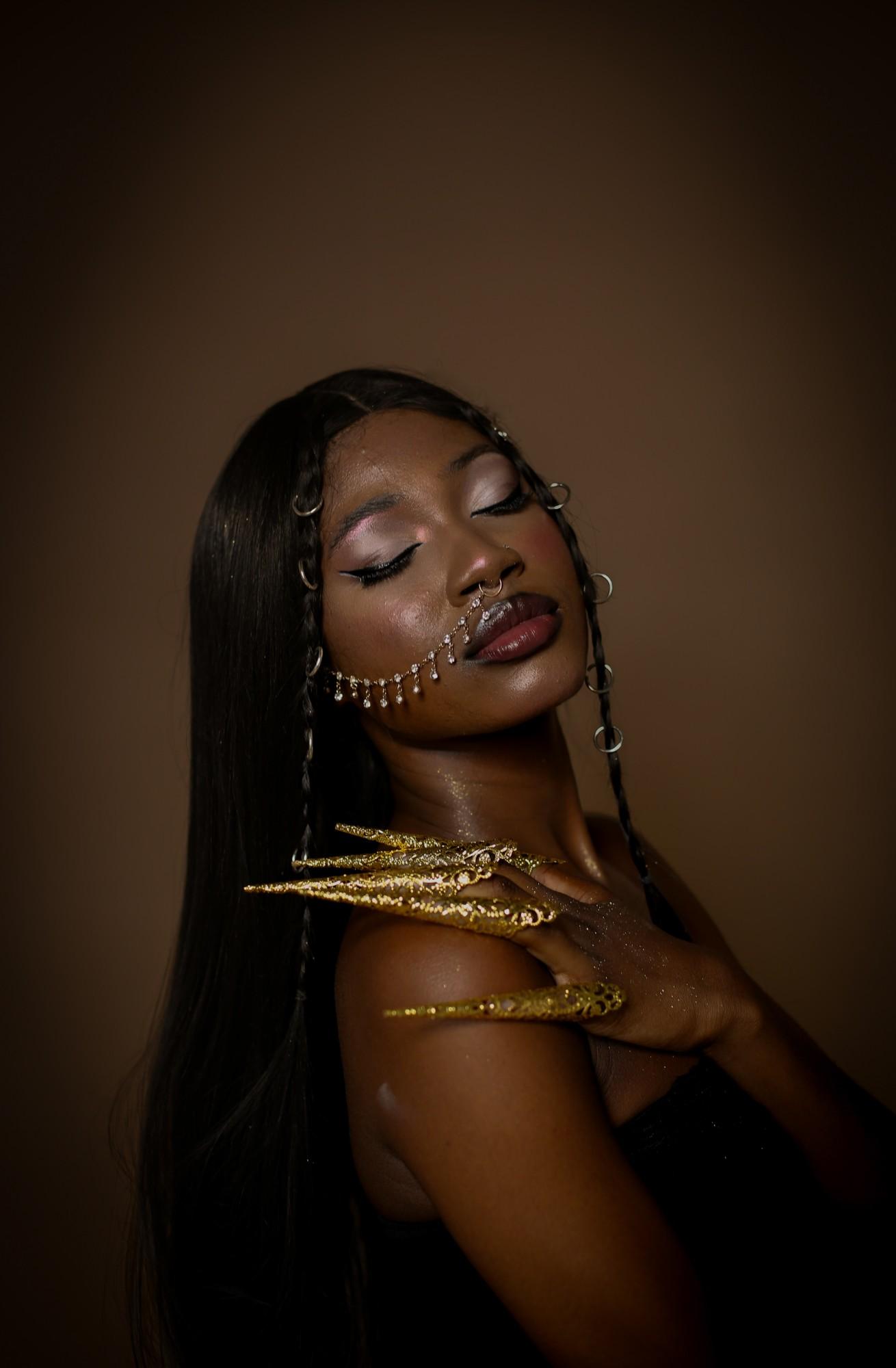 Studio portrait on a brown backdrop, eyes closed and head tilted back, gold filigree finger claws resting on one shoulder and a crystal chain across the cheek.