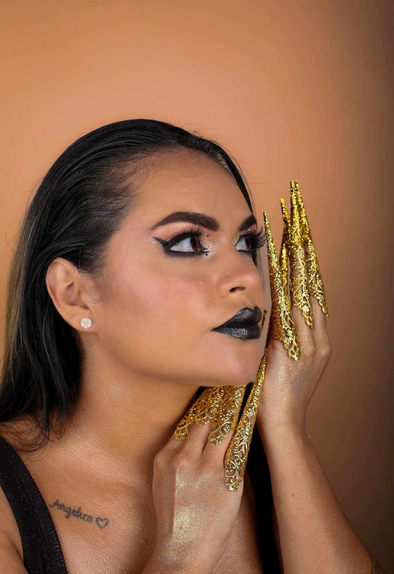 Profile portrait on an orange backdrop, looking up, black lips and winged liner, gold filigree claws on both hands framing the face.