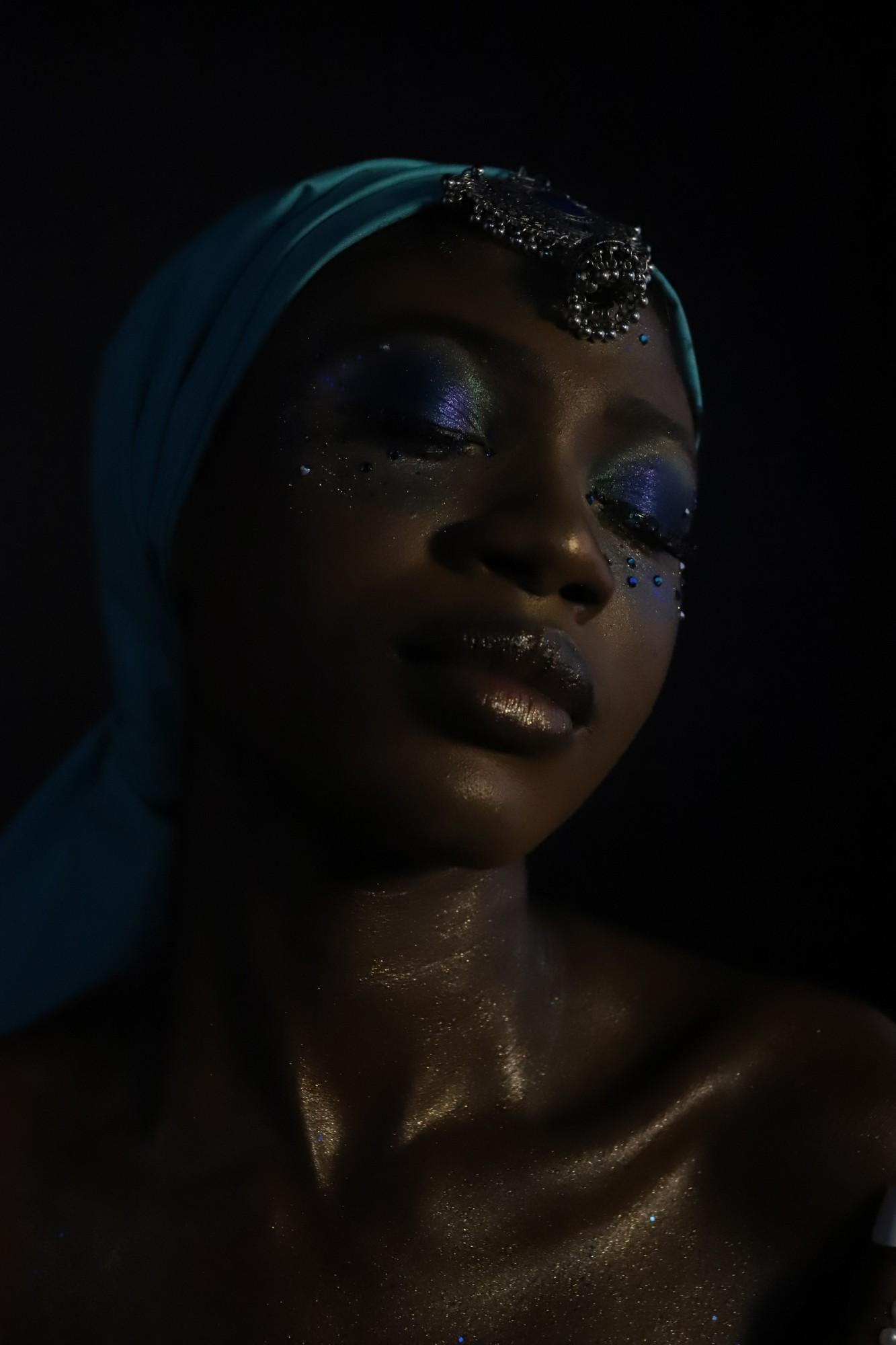 Low-key portrait on black: a teal head wrap with a silver ornament, violet shimmer on the eyelids and gold glitter catching the light on the skin.