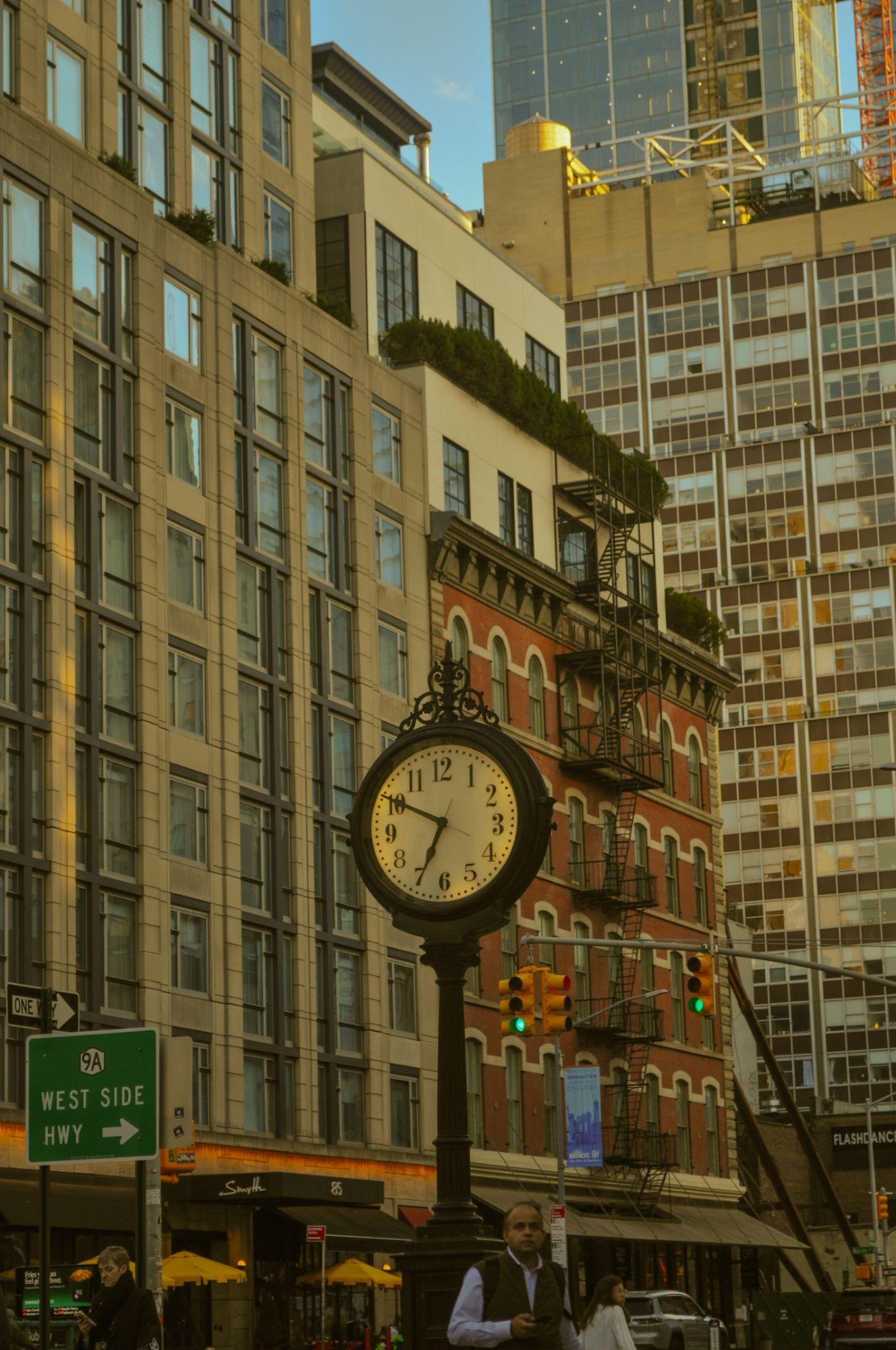 A cast-iron street clock in front of brick and glass towers in late sun, a West Side Highway sign at the left edge.